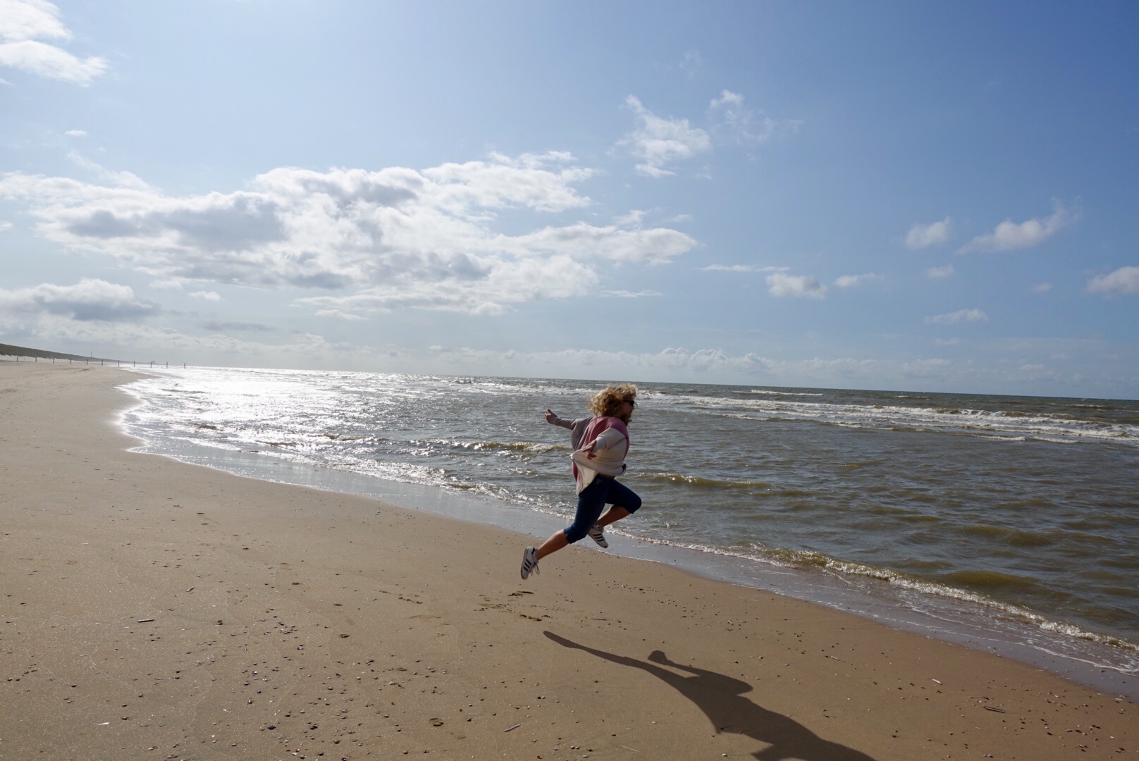 Lizzi springt ausgelassen vom Strand Richtung Meer, das von gleißendem Sonnenlicht mit Glanz überzogen wird.