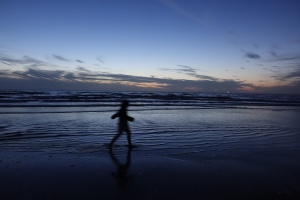 Eine schemenhafte Gestalt läuft im seichten Meerwasser vor einem von Lichtspiel und Wolken gezeichneten Himmel am Strand entlang.