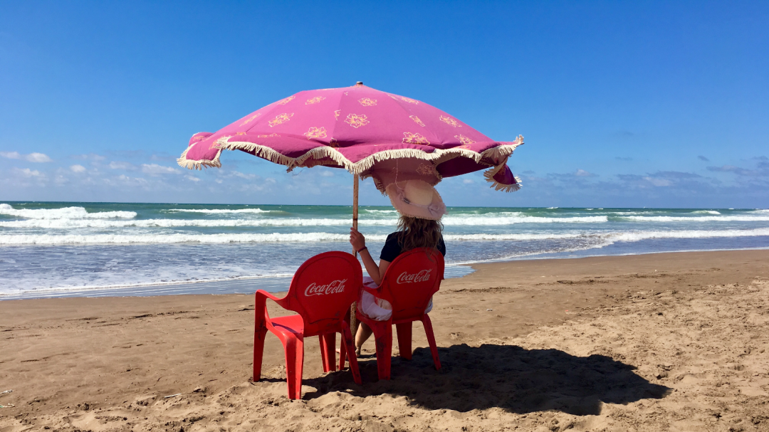 Lizzi sitzt unterm Sonnenschirm auf rotem Stuhl am Meer.