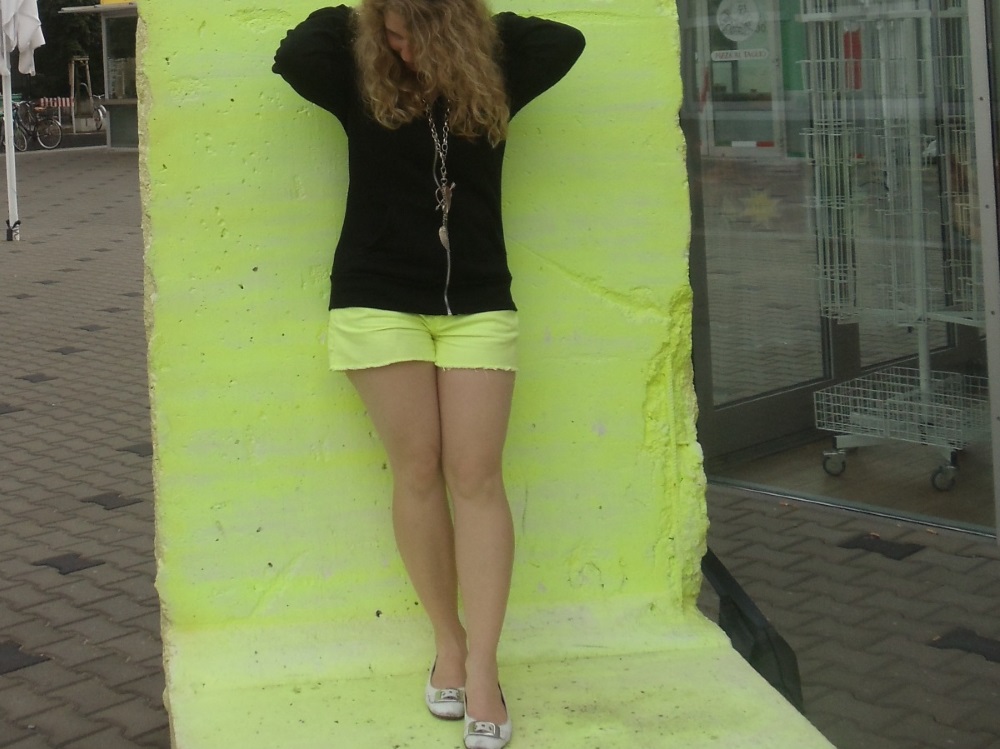 Lizzi trägt ein schwarzes Oberteil und eine leuchtendgelbe Hose. Sie steht vor einer leuchtend gelben Wand.