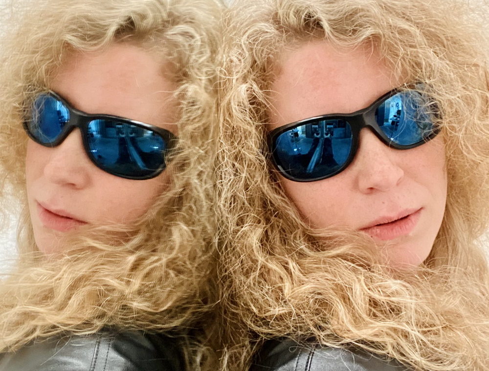 Lizzi lehnt an einem Spiegel, sodass sie doppelt zu sehen ist. Sie trägt eine schwarze Lederjacke und eine Gletschersonnenbrille. Ihre blonden Locken umrahmen ihr Gesicht.