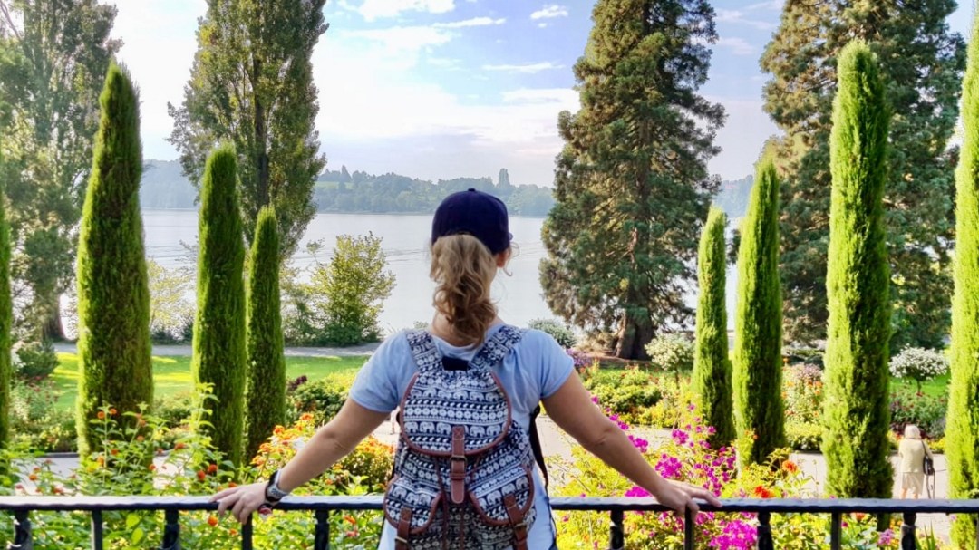 Lizzi stützt sich auf ein Geländer mit weit ausgebreiteten Armen und blickt in die Ferne. Sie trägt einen fröhlich-bunten Rucksack. Vor ihr ragen schlanke, buschige Bäume auf, im Hintergrund erstreckt sich leicht diesig der Bodensee.