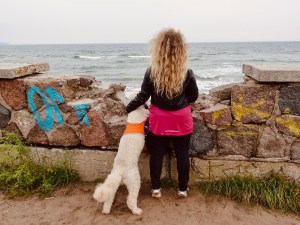 Lizzi und Blindenführhund Harry schauen über eine zerbrochene Mauer hinweg aufs dahinterliegende Meer. Harry trägt seine orangene Kenndecke, Lizzi ist in Schwarz und Pink gekleidet, die goldblonden Locken heben sich deutlich von der dunklen Lederjacke ab. Sie hat einen Arm um Harry gelegt. 