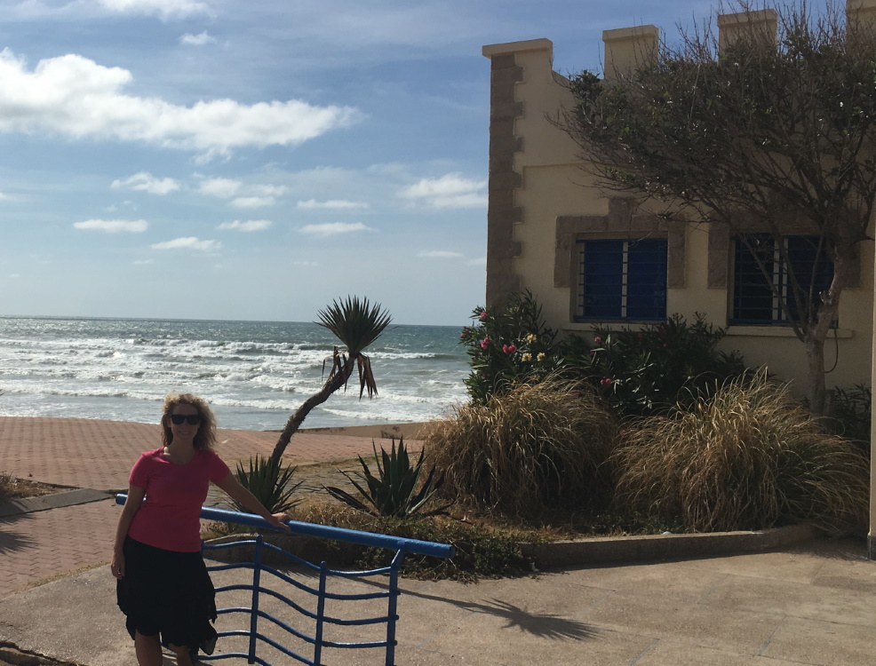 Lizzi lehnt an ein Geländer, im Hintergrund sind Strand, Meer und ein Marokkanisches Gebäude. Alles in hellem Sonnenschein.