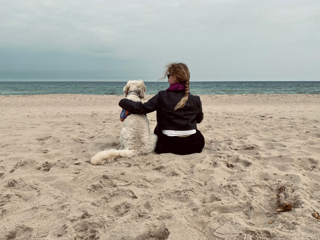 Lizzi und Harry sitzen nebeneinander im Sand, den Blick zum Meer gewandt. Lizzi hat einen Arm um den Goldendoodle gelegt.